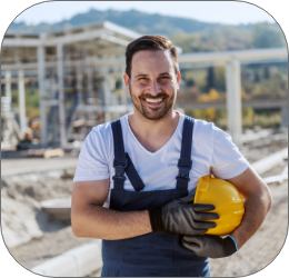 Smiling handsome unshaven caucasian worker in overall standing at refinery with helmet under armpit and looking at camera